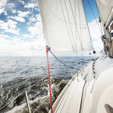 Açık bir günde açık denizde yelken açan beyaz yelkenli bir yat. Güverteden pruvaya bir manzara. Kümülüs bulutları. Ulaşım, seyahat, gezi, spor, eğlence, boş zaman aktivitesi, yarış, yelkenli yarışı