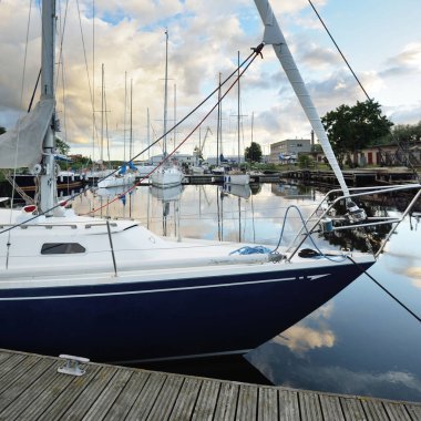 Gün batımında yat limanındaki bir rıhtıma demirlenmiş mavi yelkenli. Dramatik gökyüzü, parlayan altın bulutlar. Tatiller, ulaşım, spor, eğlence, turizm, seyahat, liman