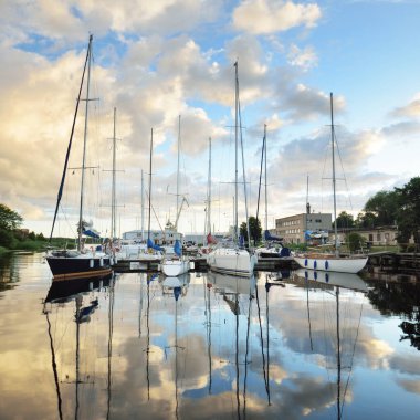 Günbatımında yat limanında demirlemiş zarif ve modern yelkenliler. Dramatik gökyüzü, parlayan altın bulutlar. Tatiller, ulaşım, spor, eğlence, turizm, seyahat, liman