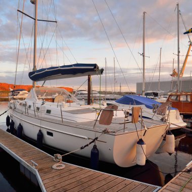 Günbatımında yat limanındaki bir rıhtıma demirlenmiş ahşap meşe güverteli modern yelkenli. Dramatik gökyüzü, parlayan altın bulutlar. Tatiller, ulaşım, spor, eğlence, turizm, seyahat, liman