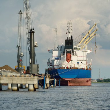 Liman terminalinde büyük bir kargo gemisi yükleniyor, arka planda vinçler var. Yük taşımacılığı, deniz taşımacılığı, lojistik, küresel iletişim, dünya çapında taşımacılık, ekonomi, ticaret, sanayi