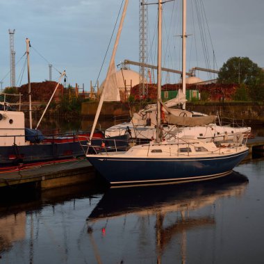 Eski römorkör ve mavi yelkenli yelkenli gün batımında yat limanındaki bir rıhtıma demirlemiş. Dramatik bir gökyüzü. Tatiller, ulaşım, spor, eğlence, turizm, seyahat, balıkçılık, liman hizmeti