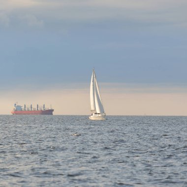 Baltık Denizi 'nde yelkenli bir yat, arkada kargo gemisi. Fırtınadan sonra açık gökyüzü, yumuşak güneş ışığı. Ulaşım, seyahat, gezi, spor, eğlence, boş zaman aktivitesi, yelkenli yarışı