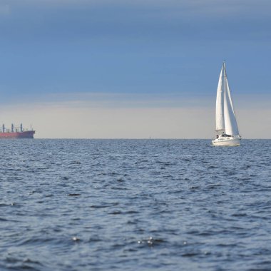 Baltık Denizi 'nde yelkenli bir yat, arkada kargo gemisi. Fırtınadan sonra açık gökyüzü, yumuşak güneş ışığı. Ulaşım, seyahat, gezi, spor, eğlence, boş zaman aktivitesi, yelkenli yarışı