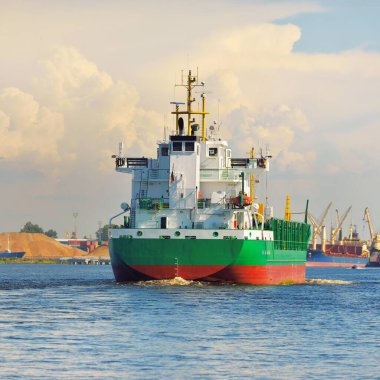 Büyük yeşil yük gemisi gün batımında kargo terminaline varacak. Arka planda gemiler ve vinçler var. Dramatik bulut manzarası. Yük taşımacılığı, küresel iletişim, endüstri, ekonomi, lojistik