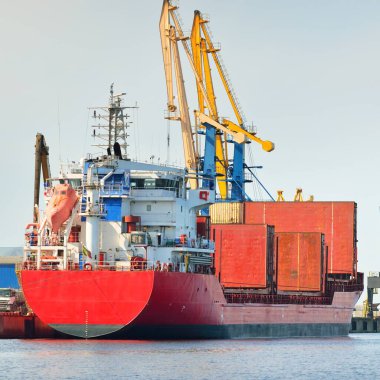 Liman terminalinde büyük bir kargo gemisi yükleniyor, arka planda vinçler var. Yük taşımacılığı, lojistik, küresel iletişim, ekonomi, ticaret, sanayi, özel ekipman
