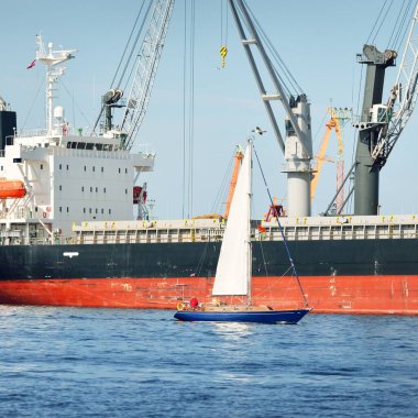 Baltık Denizi 'nde yelkenli bir yat. Kargo gemisi arka plandaki iskele terminaline yükleniyor. Ulaşım, seyahat, gezinti, spor, eğlence, eğlence etkinliği, iş, ekonomi, sanayi