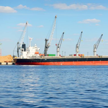 Liman terminalinde büyük bir kargo gemisi yükleniyor, arka planda vinçler var. Yük taşımacılığı, lojistik, küresel iletişim, ekonomi, ticaret, sanayi, özel ekipman