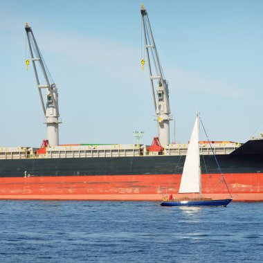Baltık Denizi 'nde yelkenli bir yat. Kargo gemisi arka plandaki iskele terminaline yükleniyor. Ulaşım, seyahat, gezinti, spor, eğlence, eğlence etkinliği, iş, ekonomi, sanayi