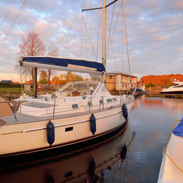 Современная парусная лодка с деревянной палубой из тика пришвартована к пирсу в яхтенной пристани на закате. Драматическое небо, светящиеся золотые облака. Отдых, транспорт, спорт, отдых, туризм, круизы, портовые услуги