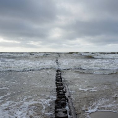 Kumlu sahilden Baltık Denizi manzarası (kum tepeleri). Dramatik gökyüzü, karanlık, parlak bulutlar. Dalgalar, su sıçramaları. Balıkçı tekneleri için eski iskele. Deniz Burnu. Bahar, iklim, doğa