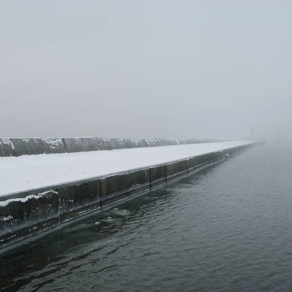 Kumlu sahilden Baltık Denizi 'nin panoramik manzarası. Deniz fenerine dolaş, dalgalar. Yoğun beyaz sis, sis, kar. Dalgalar, su sıçramaları. Deniz Burnu. Tek renkli kış manzarası