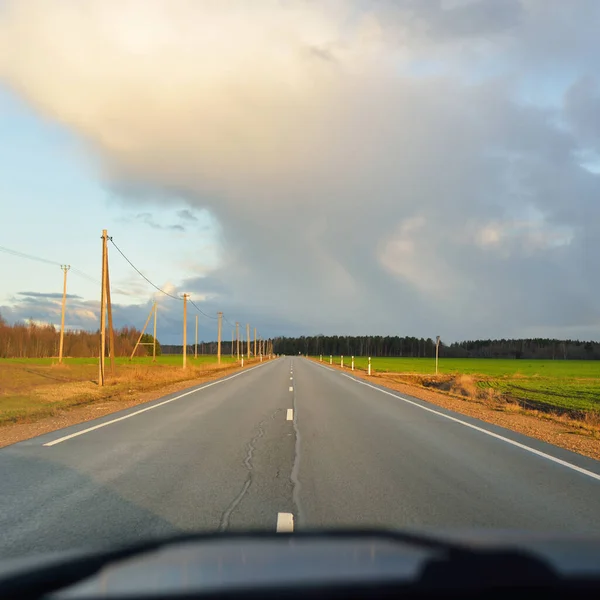 Gün batımında tarla ve orman boyunca asfalt yol (karayolu). Arabadan görüntü. Turizm, tatiller, uzak yerler, lojistik kavramlar