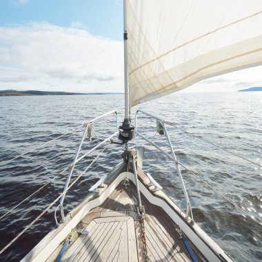 Kayalık göl kıyılarında seyir halindeki ahşap meşe güvertesiyle donatılmış modern bir yat. İskoçya, İngiltere. Güverteden pruvaya, direğe, yelkenlere bakın. Seyahat tutkusu, eğlence aktivitesi, spor, yelkenli yarışı, deniz yolculuğu.