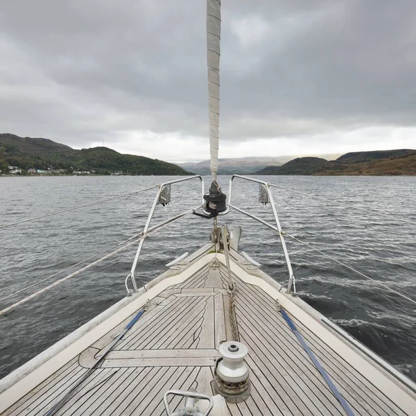 Sloop rigged modern yacht with wooden teak deck sailing near the rocky lake shores. Scotland, UK. View from the deck to the bow, mast, sails. Wanderlust, leisure activity, sport, regatta, cruise