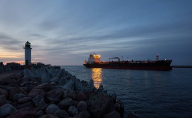 Büyük petrol kimyasal tanker gemisi gün batımında kargo limanına varacak. Dalgalar, deniz fenerine gezinti. Baltık Denizi, Ventspils, Letonya. Ulaşım, lojistik tema. Panoramik görünüm