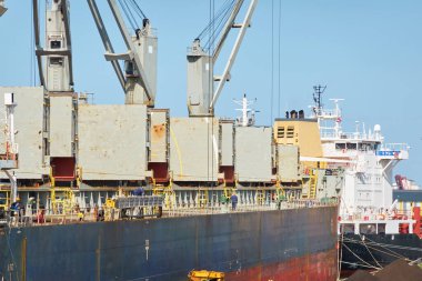 Liman terminalinde büyük bir kargo gemisi yükleniyor, arka planda vinçler var. Yük taşımacılığı, deniz taşımacılığı, lojistik, küresel iletişim, dünya çapında taşımacılık, ekonomi, ticaret, sanayi