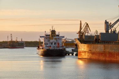 Liman terminaline büyük bir kargo gemisi yükleniyor. Gün batımı. Yük taşımacılığı, deniz taşımacılığı, lojistik, küresel iletişim, dünya çapında taşımacılık, ekonomi, ticaret, sanayi