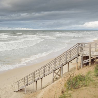 Fırtınadan sonra Baltık kıyısı (plaj). Modern tahta kaldırım (tahta kaldırım). Kum tepeleri. Dramatik gökyüzü, dalgalar ve su sıçramaları. Doğa, ekoloji, eko-turizm, yürüyüş, eğlence kavramları