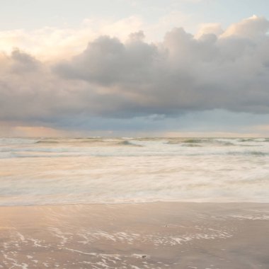 Gün batımında Baltık Denizi. Dramatik gökyüzü, mavi ve pembe parlayan bulutlar, altın güneş ışığı. Dalgalar, su sıçratma. Resimli manzara, deniz manzarası, doğa. Panoramik görünüm, uzun pozlama