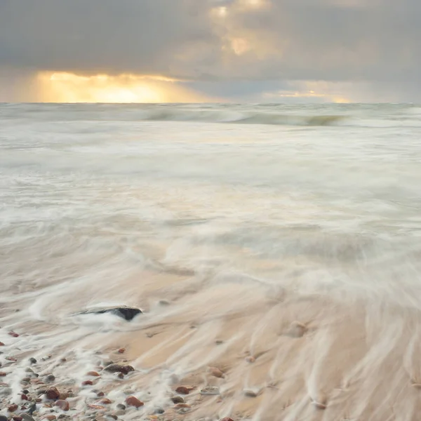 Gün batımında Baltık Denizi. Dramatik gökyüzü, mavi ve pembe parlayan bulutlar, altın güneş ışığı. Dalgalar, su sıçratma. Resimli manzara, deniz manzarası, doğa. Panoramik görünüm, uzun pozlama