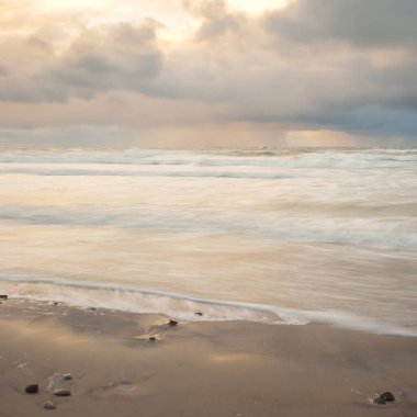 Gün batımında Baltık Denizi. Dramatik gökyüzü, mavi ve pembe parlayan bulutlar, altın güneş ışığı. Dalgalar, su sıçratma. Resimli manzara, deniz manzarası, doğa. Panoramik görünüm, uzun pozlama
