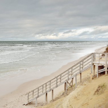 Fırtınadan sonra Baltık kıyısı (plaj). Modern tahta kaldırım (tahta kaldırım). Kum tepeleri. Dramatik gökyüzü, dalgalar ve su sıçramaları. Doğa, ekoloji, eko-turizm, yürüyüş, eğlence kavramları