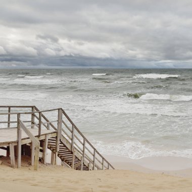 Fırtınadan sonra Baltık kıyısı (plaj). Modern tahta kaldırım (tahta kaldırım). Kum tepeleri. Dramatik gökyüzü, dalgalar ve su sıçramaları. Doğa, ekoloji, eko-turizm, yürüyüş, eğlence kavramları