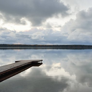 Gün batımında Orman Nehri (Lake). Ahşap iskele. Dramatik gökyüzü, karanlık, parlak bulutlar. Kristal berrak suda simetri yansımaları. Panoramik sonbahar manzarası. Doğa, ekoloji, ekoloji, ekolojik tatil beldesi