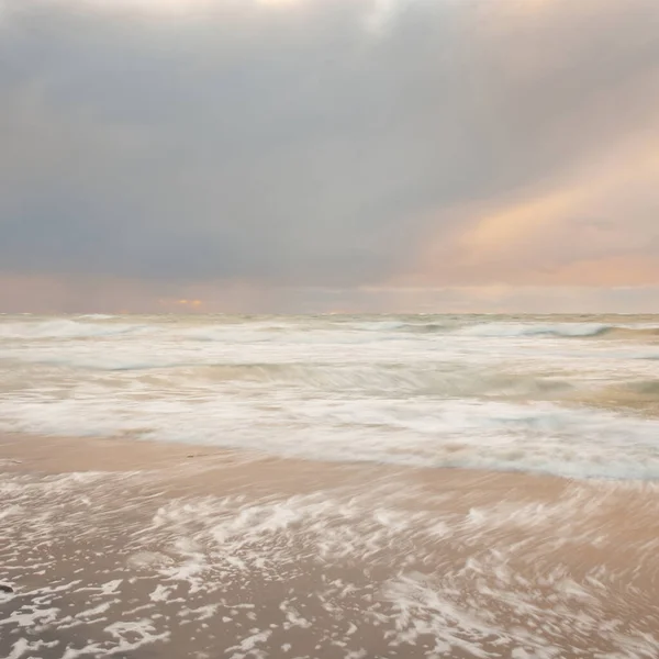 Gün batımında Baltık Denizi. Dramatik gökyüzü, mavi ve pembe parlayan bulutlar, altın güneş ışığı. Dalgalar, su sıçratma. Resimli manzara, deniz manzarası, doğa. Panoramik görünüm, uzun pozlama
