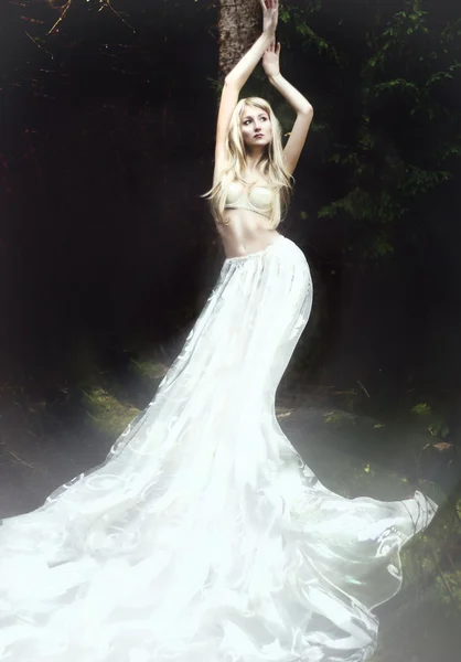 Blond anděl v dlouhé bílé sukni, stojící v temném lese — Stock fotografie
