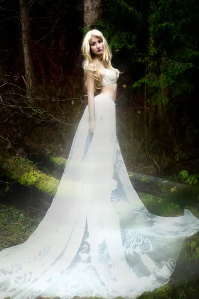 Blonde bride angel in long white skirt standing in dark forest