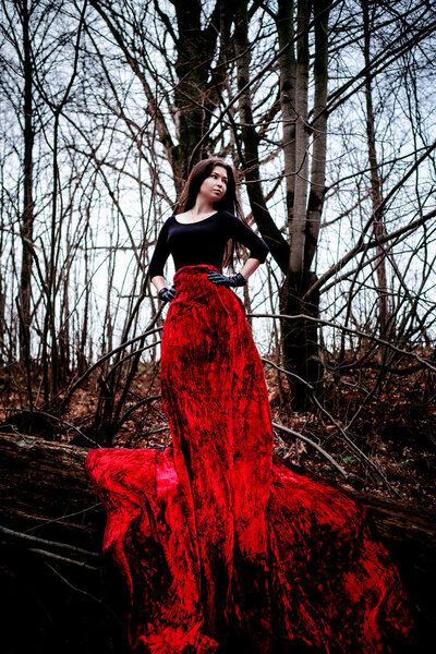 Mysterious woman or witch in long red dress standing in dark forest