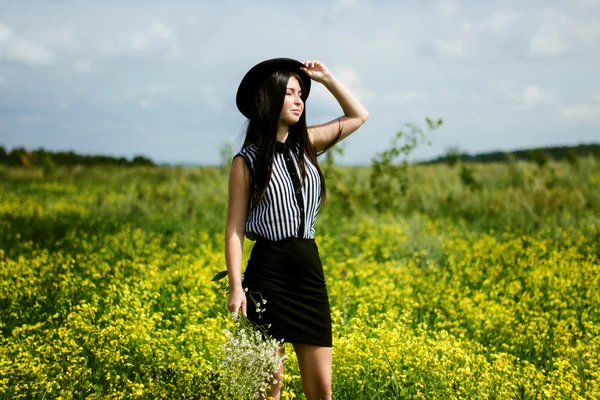 戴着黑帽子站在阳光花黄色字段上的时尚女人 — 图库照片