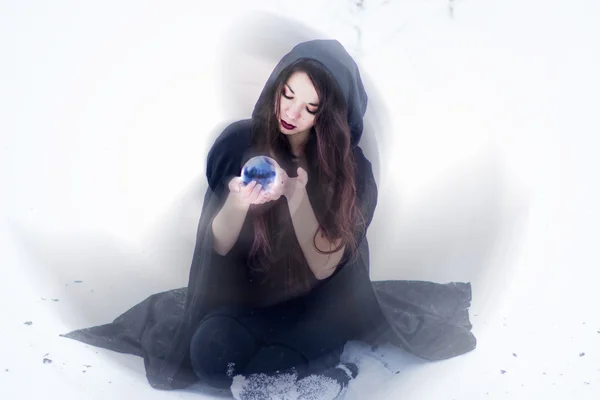 Witch or woman doing magic in black cloak with glass ball in white snow forest — Stock Photo, Image