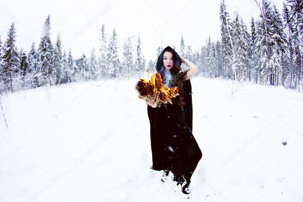 Witch or woman in black cloak with fire ball in white snow forest