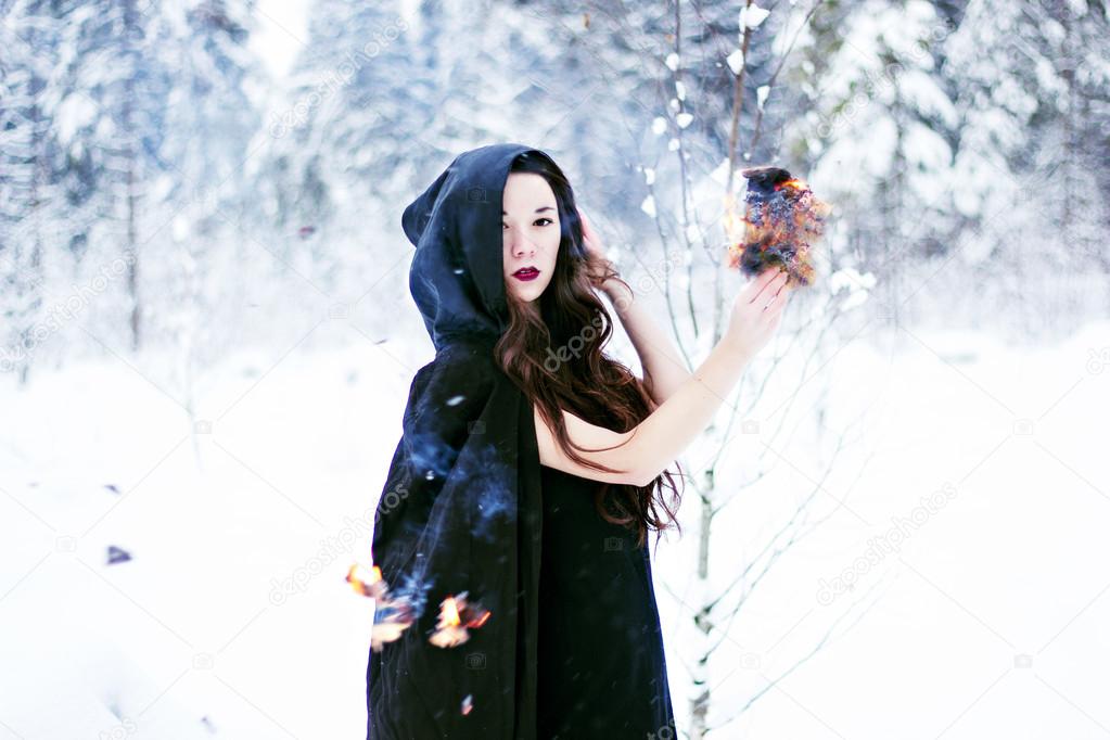 Witch or woman in black cloak with fire ball in white snow forest