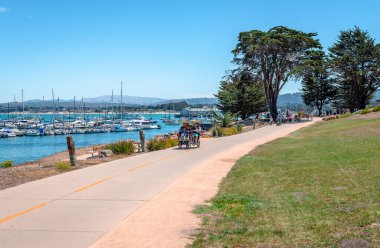 Monterey, CA / USA - July 18 2015: People enjoy a walk in Monterey Bay Coastal Recreation Trail on a sunny day, with the Old Fisherman's Wharf and many boats in the background.