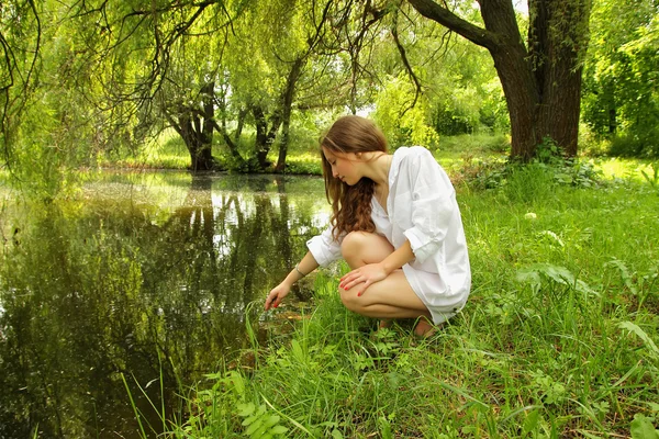 Chica joven con el pelo largo sentado cerca del lago a la sombra —  Fotos de Stock