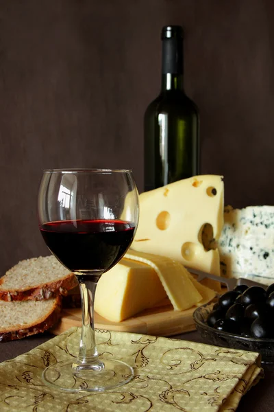 Bardak kırmızı şarap, şişe ve birkaç çeşit peynir, cam üzerinde odak — Stok fotoğraf