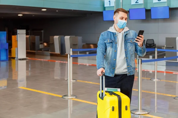 Man in medical face mask using smartphone behind check in counter at airport interior during covid lockdown. Urban casual traveler holds mobile phone passport suitcase luggage. Man in airport