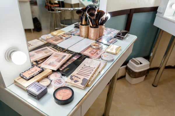 Mesa de espaço de trabalho de maquiador profissional com paletas de sombra, produtos cosméticos e pincéis. Cosméticos de diferentes marcas maquiador na mesa com espelho e lâmpadas no salão de beleza — Fotografia de Stock