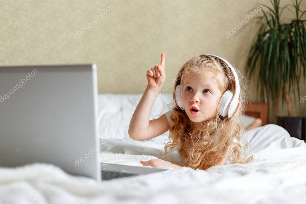 Caucasian blonde child girl use laptop in headphones for doing homework. Girl lying on bed Discuss idea With interest Curiosity via video call. Distance learning E-learning at home for children