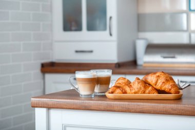 Kahverengi mutfak masasında kahvaltıda kruvasan ve sütlü iki kahve. Şeffaf bardaklarda bir çift cappuccino ve Fransız kruvasanları romantik bir kahvaltı için güvertede.