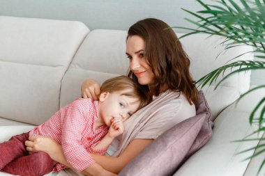 Bebek, annesinin göğsünde, iç kısımdaki kanepede uzanmış parmağını emiyor. Gülümseyen anne ve kızı pijamalarıyla kanepede uzanmış birbirlerine sarılıyorlar.