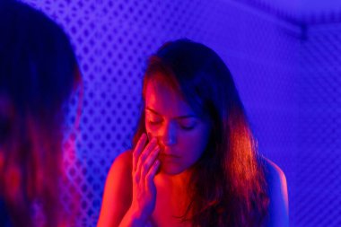 Geceleri banyoda depresyonda olan üzgün, yorgun bir kadının ayna yansıması. İntihar yalnızlığı, hastalık bağımlılığı ve akıl sağlığı kavramı. Neon Mavi ve Kırmızı Işık