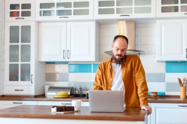 Adam online iş eğitimi veya alışveriş için dizüstü bilgisayar kullanıyor. Cesur Sakallı Kafkaslı Adam Freelancer 'ın portresi mutfaktaki ofisini kullanıyor. Evdeki çalışma odası ahşap masa. Evden çalışan sakin genç adam.. 