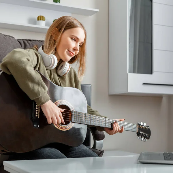 Genç kız yakın çekim portresi evde gitar çalmayı öğrenirken çevrimiçi dersleri kullanıyor. Uzaktan hobi müzik eğitimi akustik gitar. Genç bir kadın dizüstü bilgisayarla evde akustik gitar çalıyor.