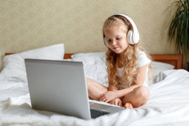 Kafkasyalı sarışın kız video veya çizgi film izlemek için kulaklıkla dizüstü bilgisayar kullanıyor. Kız video aracılığıyla yatak derslerine katılıyor. Çocuklar için evde uzaktan öğrenme E-öğrenimi
