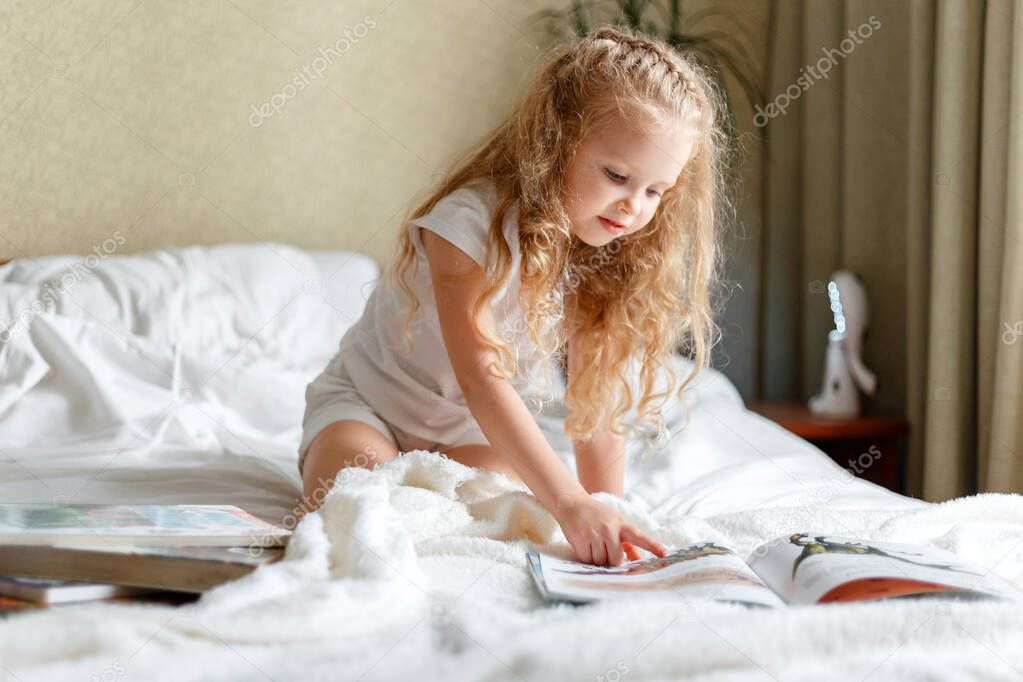 Beautiful little girl Child read book on bed in morning. Little blond curly girl Read book in bedroom sunny Childrens room at home. Caucasian Girl doing homework study or imagine on bed